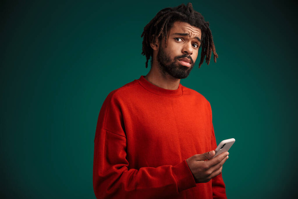 Portrait of unhappy african american man using cellphone and looking at camera isolated over green background - Fotografie, Obrázek