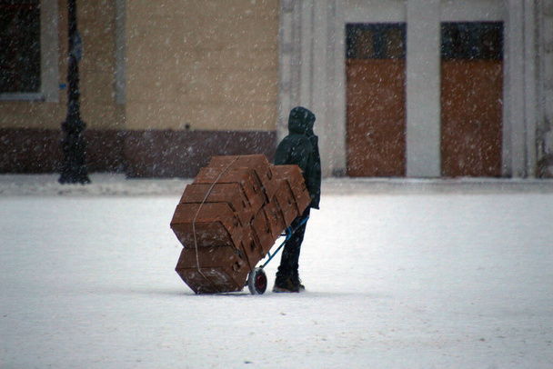 Człowiek niosący dużo kartonowych pudełek na wózku na śnieżnej drodze podczas burzy śnieżnej - Zdjęcie, obraz