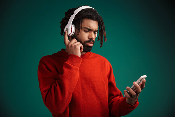 Portrait of serious african american man using cellphone and wireless headphones isolated over green background - Foto, afbeelding
