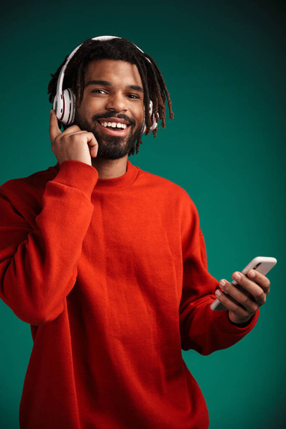 Portrait of laughing african american man using cellphone and wireless headphones isolated over green background - Foto, imagen