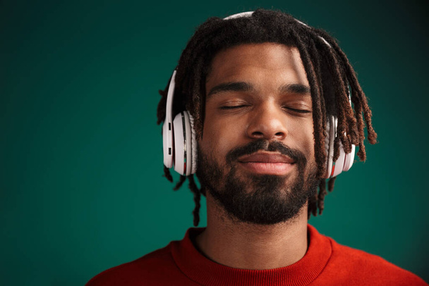 Portrait of pleased african american man smiling and using wireless headphones isolated over green background - Foto, Bild