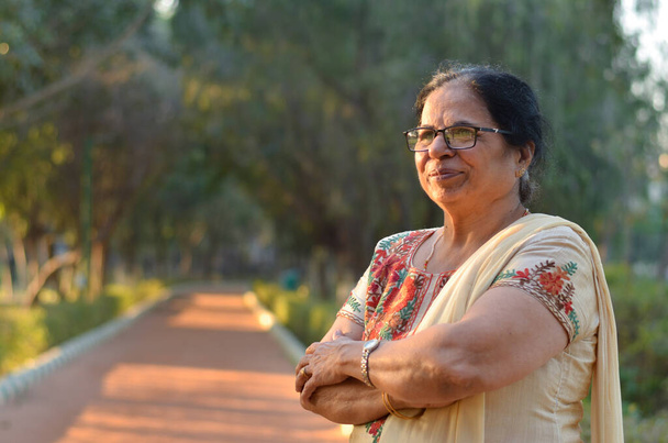 Έξυπνη και σίγουρος ανώτερος βόρεια ινδική γυναίκα στέκεται, σκέψης και κοιτάζοντας μακριά με τα χέρια σταυρωμένα / διπλωμένα σε ένα πάρκο φορώντας ένα off λευκό κοστούμι salwar kameez punjabi το καλοκαίρι στο Νέο Δελχί, Ινδία - Φωτογραφία, εικόνα