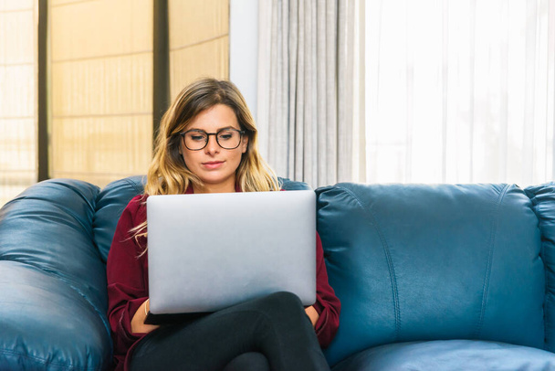 Een glimlachende blanke vrouw die op haar blauwe bank zit met haar laptop. Ze draagt een bril.. - Foto, afbeelding