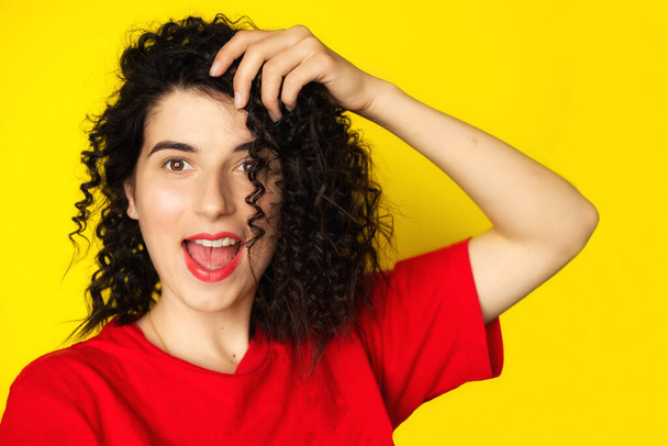 Charming young girl with curly black hair in a red T-shirt on a yellow background. Portrait photography. Emotions, facial expressions. Curls, Brunette. Red lipstick - Photo, Image