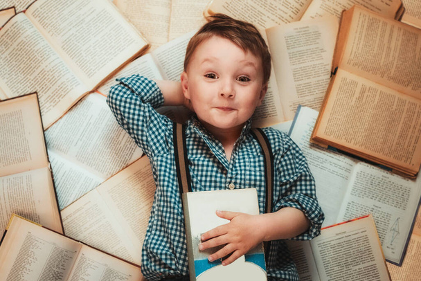на книгах лежит маленький мальчик в клетчатой рубашке и синих брюках с подтяжками. Концепция книжного магазина. Книги это знание. Вид сверху
 - Фото, изображение