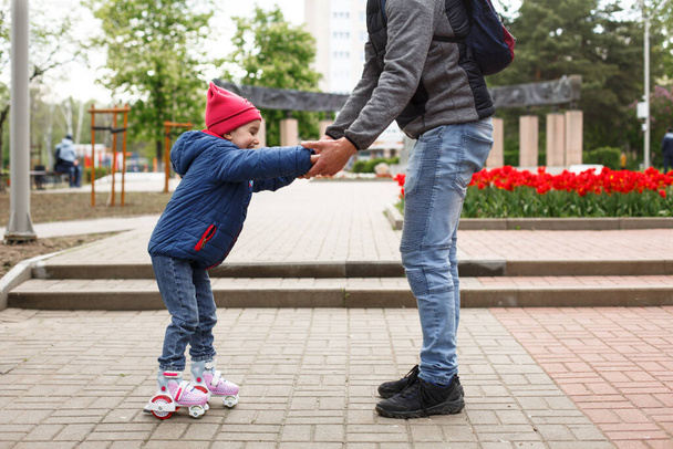 Dad teaches his little daughter to ride four-wheeled roller skates. The father supports the child by the hands, feet of the father and the child in skates. Focus on the rollers. Father's day, family weekend, place for text - Zdjęcie, obraz