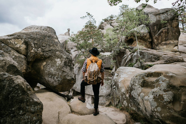 Um homem com uma jaqueta laranja e chapéu fica em uma rocha e olha para a distância. Conceito de viagem de montanha
 - Foto, Imagem