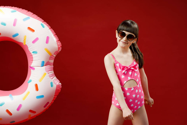 Isolated shot of charming little girl in sunglasses posing in studio with rubber ring going to have swim in sea during vacations. Pretty female child holding swimming inflatable circle - Photo, image