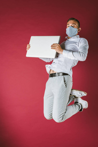 jeune homme en chemise blanche et masque sur son visage sauta, pleine longueur avec un ordinateur dans les mains, sur un fond rouge. Pandimie, isolement
 - Photo, image