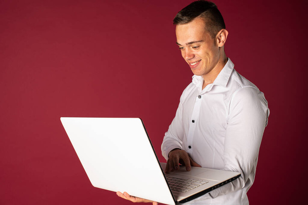 Pewny siebie ekspert od interesów. Pewny siebie młody przystojny mężczyzna w koszuli trzyma laptopa i uśmiecha się stojąc na czerwonym tle - Zdjęcie, obraz