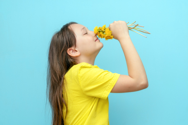 Mavi bir arka planda elinde çiçekli karahindiba olan bir kız çocuğunun portresi. Genç kız çiçekleri kokluyor ve sevinçle gözlerini kapıyor. Çiçeklere karşı sezonluk polen alerjisi kavramı. Zaman. - Fotoğraf, Görsel