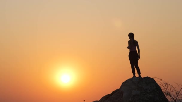 Η σιλουέτα μιας γυναίκας πεζοπόρου που στέκεται μόνη της σε μια μεγάλη πέτρα το ηλιοβασίλεμα στα βουνά. Γυναίκα τουρίστρια απολαμβάνοντας θέα στο ψηλό βράχο στη φύση το βράδυ. Τουρισμός, ταξίδια και υγιεινός τρόπος ζωής. - Πλάνα, βίντεο