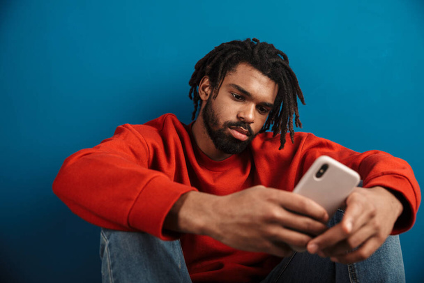 Close up portrait of a young african man wearing pullover sitting isolated over blue background, using mobile phone - Foto, afbeelding