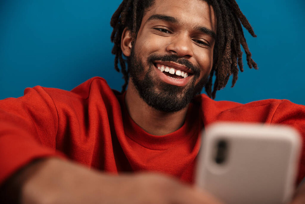 Close up portrait of a smiling young african man wearing pullover sitting isolated over blue background, using mobile phone - Φωτογραφία, εικόνα