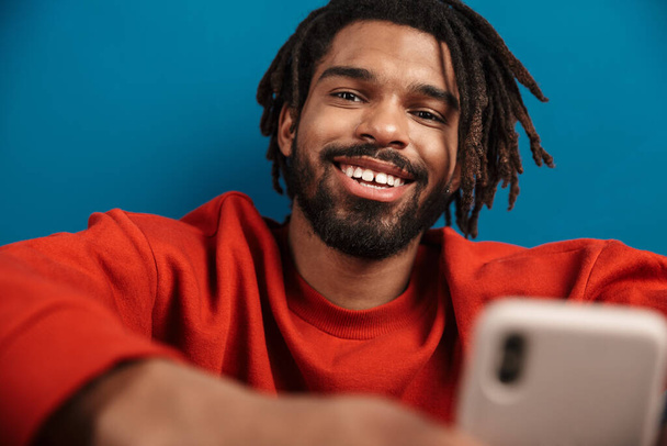 Close up portrait of a smiling young african man wearing pullover sitting isolated over blue background, using mobile phone - 写真・画像
