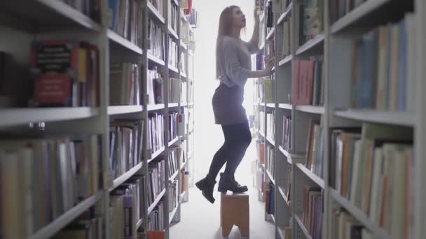 Piękna blondynka student dziewczyna biorąc książkę z półki w bibliotece uniwersyteckiej. - Materiał filmowy, wideo