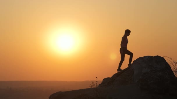 Silhouet van een vrouwelijke wandelaar die alleen klimt op grote steen bij zonsondergang in de bergen. Vrouwelijke toerist die 's avonds haar handen opsteekt op hoge rotsen. Toerisme, reizen en een gezonde levensstijl. - Video