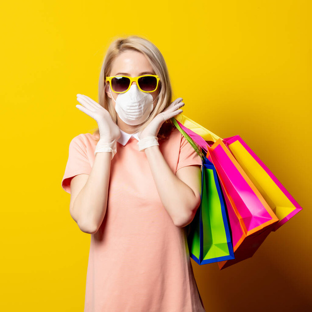 Blonde girl in face mask and pink dress hold shopping bags on yellow background. - Zdjęcie, obraz