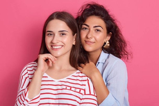 Two happy young female friends standing against rosy background, wearing stylish casuals and looking at camera, ladies expressing positive emotions, girl with straight hair keeping hand under chin. - Zdjęcie, obraz