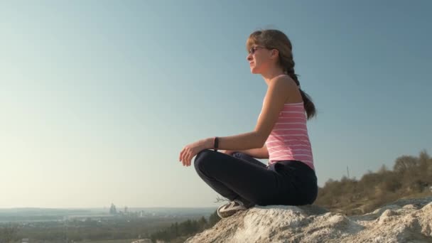 Молодая расслабленная женщина сидит на открытом воздухе на большом камне наслаждаясь теплым летним днем. Девушка медитирует и отдыхает на природе на закате. - Кадры, видео