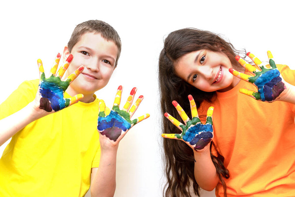 a boy and a girl of 10 years old look at the camera and show their palms in paints. Rainbow on the hands. The concept of a happy childhood and childrens day. Space for text - Photo, Image