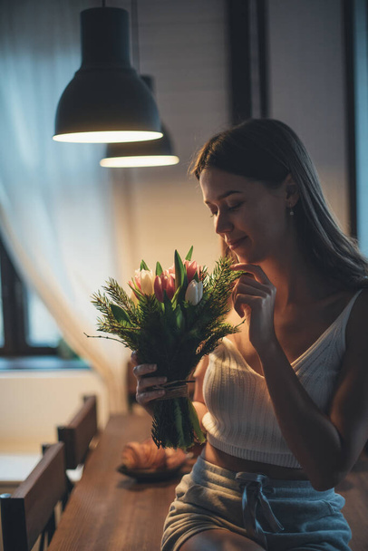 Nuoren naisen muotokuva kukkakimpun kanssa. Kaunis tyttö istuu pöydällä illalla valossa lamput tulppaanit käsissään. Aistillinen muotokuva hillityssä valossa - Valokuva, kuva