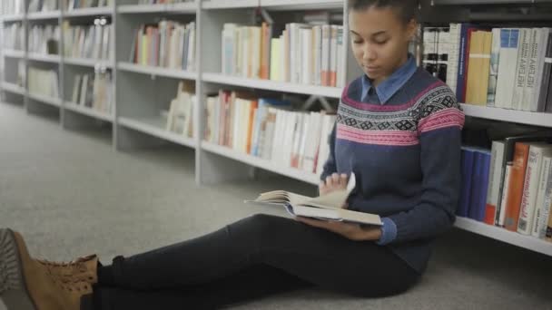 Jonge Afro-Amerikaanse vrouw leest boek zittend op de vloer in de universiteitsbibliotheek. - Video