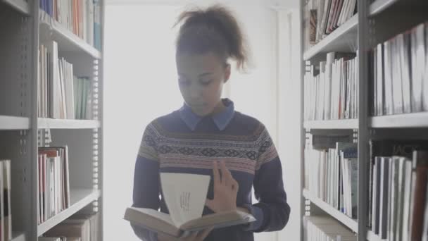 Genç kıvırcık saçlı Afrikalı Amerikalı kadın kütüphanedeki kitaplıkların arasında yürürken kitap okuyor.. - Video, Çekim