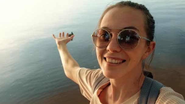 Sahilde güneş gözlüklü mutlu genç bir kadın, sakin nehir sürüsüne karşı. - Video, Çekim