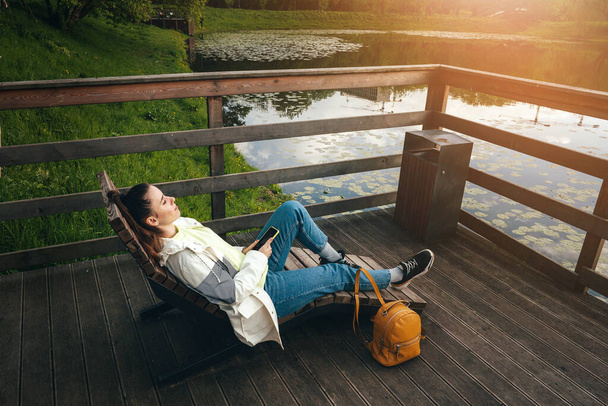 menina encontra-se em um banco de madeira perto do lago. telefone em hands.sunlight
 - Foto, Imagem