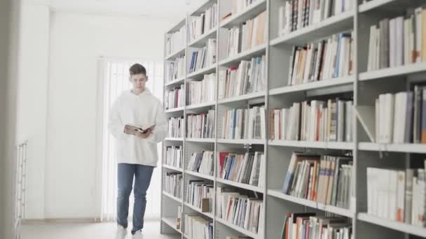 Guapo joven estudiante caminando entre estante de libros con libro en las manos
 - Imágenes, Vídeo