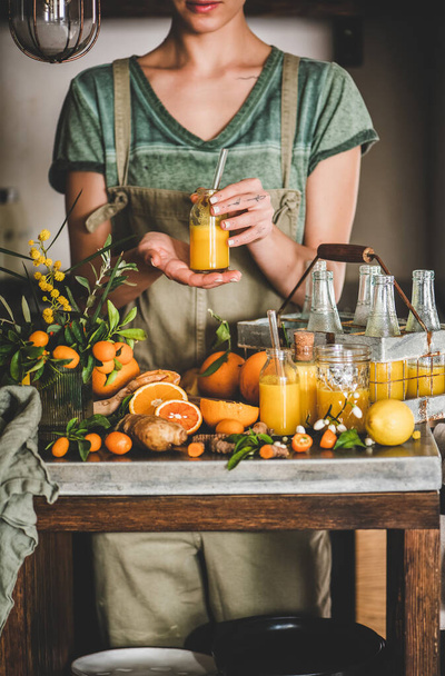 Immune boosting vitamin health defending drink. Young woman in apron holding homemade fresh turmeric, ginger, citrus juice shot in bottle in hands in kitchen. Pure vegan Immunity system booster - Foto, Imagen