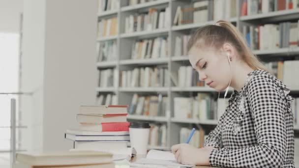 Studentessa concentrata con cuffie che si prepara per l'esame e scrive appunti mentre siede a tavola presso la biblioteca universitaria - Filmati, video