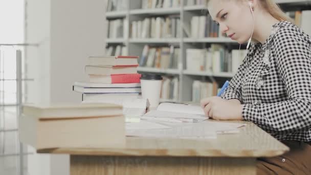 Dolly πλάνο του μαθητή κορίτσι με ακουστικά προετοιμασία για τις εξετάσεις και γράφοντας σημειώσεις, ενώ κάθεται στο τραπέζι στη βιβλιοθήκη του πανεπιστημίου - Πλάνα, βίντεο