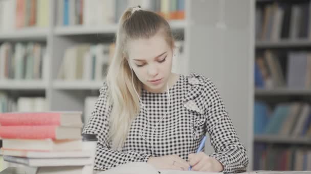 Portret van een studente met koptelefoon die zich voorbereidt op het examen en notities schrijft terwijl ze aan tafel zit in de universiteitsbibliotheek - Video