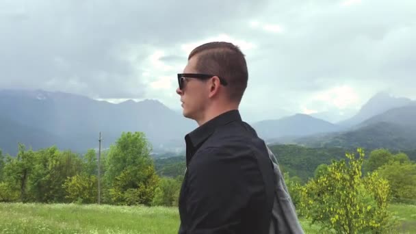 όμορφος άντρας με γυαλιά ηλίου και casual ρούχα που περπατά ανάμεσα στα βουνά την καλοκαιρινή μέρα   - Πλάνα, βίντεο