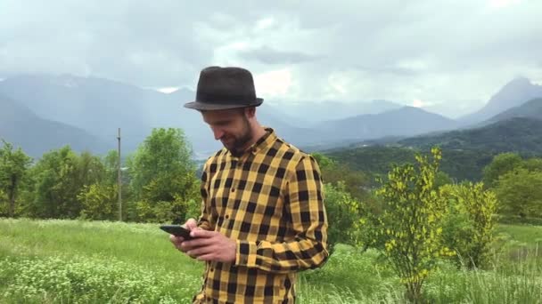 homem bonito em roupas casuais usando smartphone durante a caminhada entre as montanhas no dia de verão
 - Filmagem, Vídeo