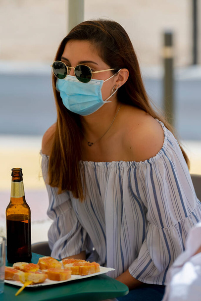 Νεαρή γυναίκα που φοράει γυαλιά ηλίου και μια χειρουργική μάσκα απολαμβάνοντας ένα σνακ και μια μπύρα κάθεται σε μια υπαίθρια βεράντα μπαρ. Νέα έννοια τρόπου ζωής. - Φωτογραφία, εικόνα