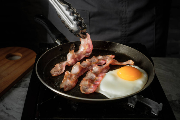 Egg and bacon cooking on pan for tasty traditional American breakfast on kitchen black background closeup - Photo, Image