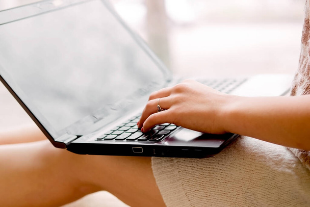 chica en un suéter de punto ligero se sienta cerca de la ventana y sostiene un ordenador portátil. Las manos femeninas imprimen en un cuaderno, trabajo remoto en hom - Foto, Imagen