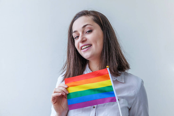 Красивая кавказская лесбиянка с радужным флагом ЛГБТ на белом фоне выглядит счастливой и взволнованной. Портрет молодой женщины гей-прайд. Равные права на концепцию сообщества lgbtq - Фото, изображение