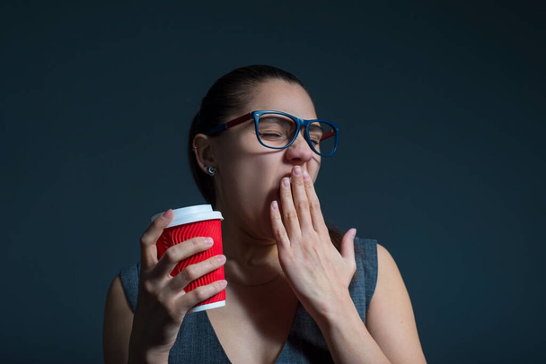Portrait of a tired yawning girl, covers her hand. With glasses, a girl drinks coffee from a paper red glass. Studio photo on a gray background. Business office style, girl tired drinks coffee, yawns. Fatigue of office workers. - Photo, Image