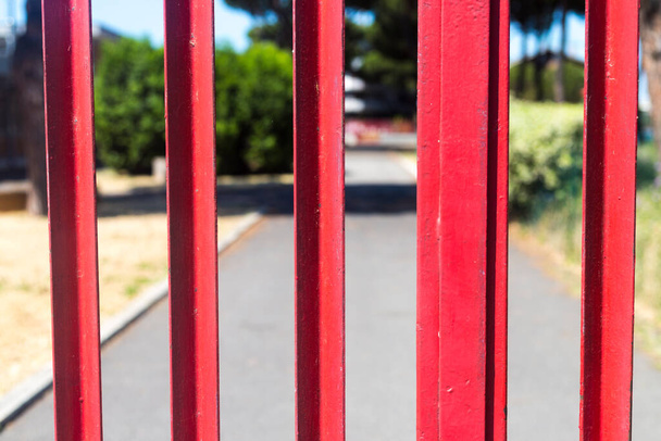 Closeup of closed red gate. School park closed for visitors. Asphalt road and alley behind a metal hence. Summer sunny day. School closed till September. No people concept - Photo, Image