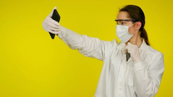 Лікар в захисній медичній масці, окулярах, рукавичках робить селфі на смартфоні
 - Фото, зображення