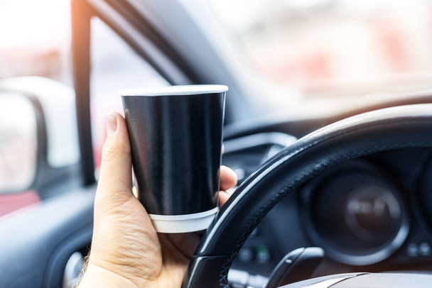 Close-up osoba ręka trzymając czarny pusty makieta papierowy kubek woth gorąca kawa lub herbata na wynos pić jazdy w mieście dżem ruchu miejskiego. Deska rozdzielcza samochodu i wschód słońca słoneczne okno tło wcześnie rano - Zdjęcie, obraz
