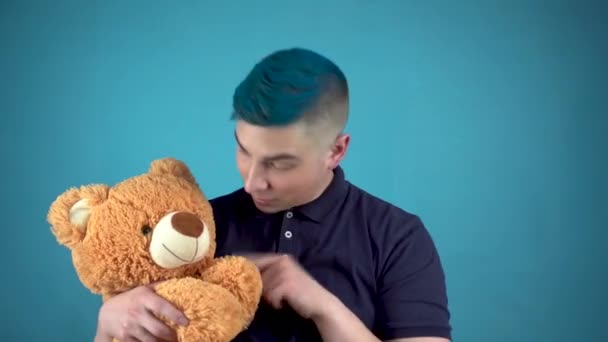 Een jonge man met blauw haar houdt een teddybeer vast. Alternatieve man zwaait met zijn hand en kijkt naar de camera op een blauwe achtergrond. - Video