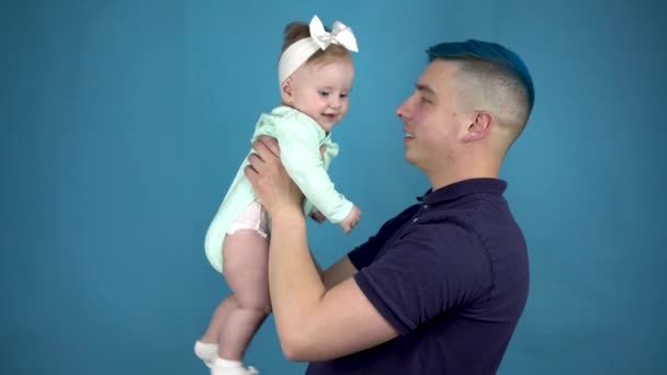 Młody ojciec z niebieskimi włosami trzyma dziecko w ramionach. Alternatywny mężczyzna z dzieckiem patrzy w kamerę na niebieskim tle. - Materiał filmowy, wideo