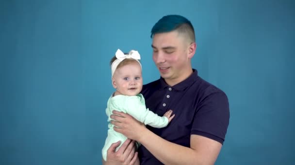 Un giovane padre con i capelli blu tiene un bambino tra le braccia. Uomo alternativo con un bambino guardare la fotocamera su uno sfondo blu. - Filmati, video