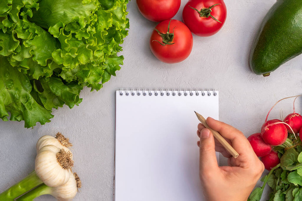 Mutfak masasındaki kağıt not defterine taze sebzelerle birlikte kalem yazan bir kadın. Alkali gıda arka planı - Fotoğraf, Görsel