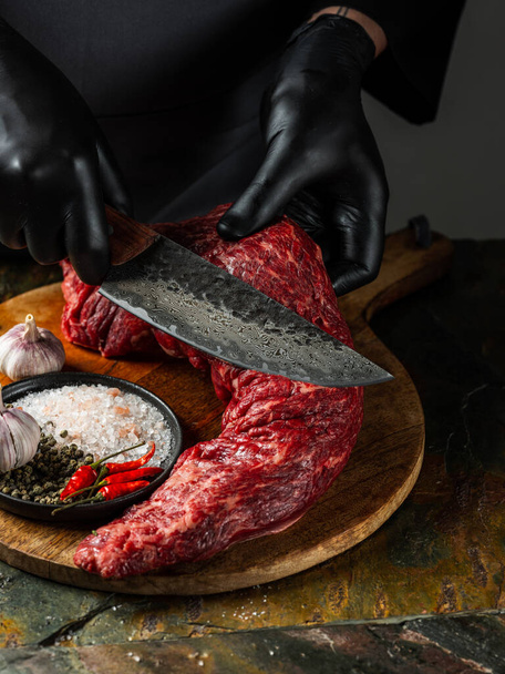 Beef fillet meat chop with big knife butcher with pepper, salt, herbs on stone table dark background. Flat top view, overview - Foto, immagini